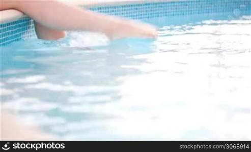 Dolly shot of young woman having fun splashing her feet in the swimming pool