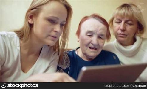 Dolly shot of young woman with tablet PC showing photos or video to her mother and elderly grandmother