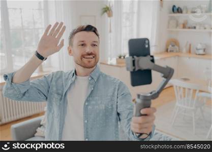 Domestic handsome guy blogger using smartphone with gimbal, waving hand in hello gesture and telling followers in social media about his life while standing in living room at home. Young handsome guy blogger making video blog on smartphone
