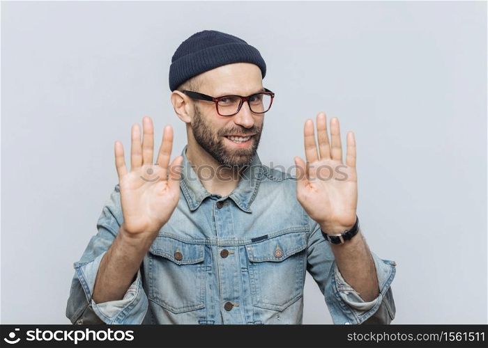 Don`t bother me! Cheerful male with pleasant appearance shows stop sign, asks not to speak with him, wears fashionable hat and glasses, isolated over grey studio background. People and denial concept