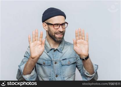 Don`t bother me! Cheerful male with pleasant appearance shows stop sign, asks not to speak with him, wears fashionable hat and glasses, isolated over grey studio background. People and denial concept