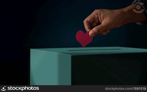 Donation Concept. Hand putting a Red Heart paper into a Donate Box. Metaphor photo. Dark tone