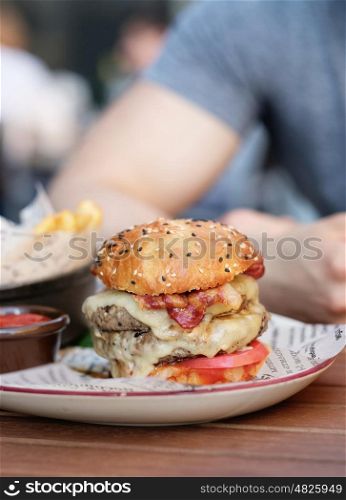 Double cheeseburger on table in outdoor restaurant with shallow depth of field. Blurred young man in background.