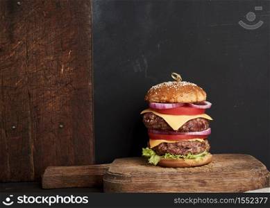double cheeseburger with tomatoes, onions, barbecue cutlet and sesame bun on an old wooden cutting board, brown background. Fast food