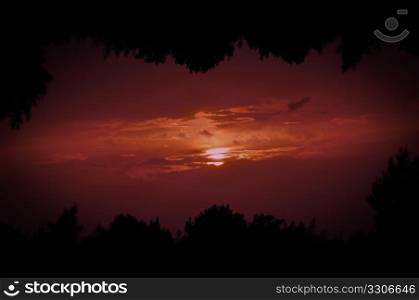 Double printed photo of setting sun behind clouds and tree silhouettes frame.