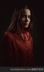 Dramatic portrait of a young beautiful woman. Lonely beautiful woman in coral shirt attentively looking at camera on dark background, low key studio shot