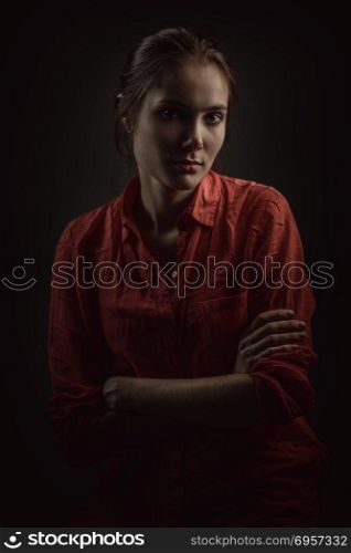Dramatic portrait of a young beautiful woman. Young beautiful woman in coral shirt with arms crossed on dark background, low key studio shot