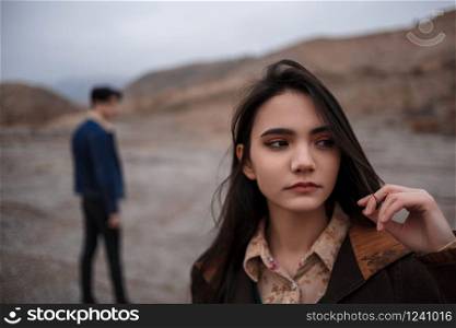 Dramatic portrait of a young brunette girl in cloudy weather. somewhere behind her, out of focus, her young lover boyfriend leaves her after break up . selective focus, small focus area.