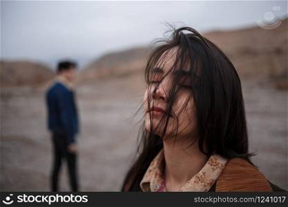 Dramatic portrait of a young brunette girl in cloudy weather. somewhere behind her, out of focus, her young lover boyfriend leaves her after break up . selective focus, small focus area.