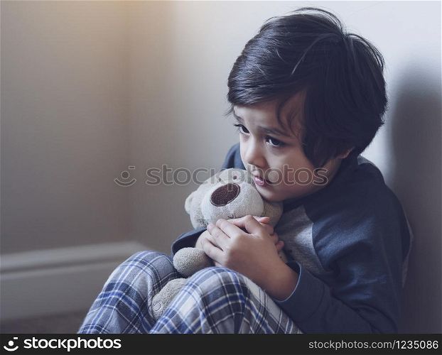 Dramatic portrait of little boy sitting on carpet cuddling teddy bear with scared face, Unhappy Child sitting alone and looking out with worrying face,Toddler boy on corner punishment sitting.
