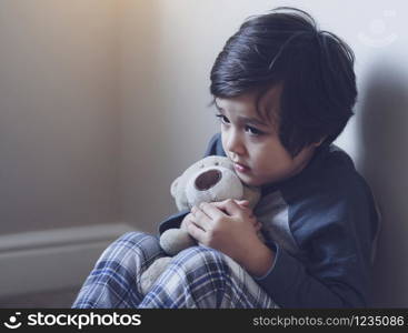 Dramatic portrait of little boy sitting on carpet cuddling teddy bear with scared face, Unhappy Child sitting alone and looking out with worrying face,Toddler boy on corner punishment sitting.