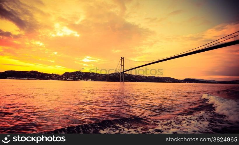 Dramatic sunset cloudy sky above a surface of the sea and long suspension bridge in Bergen, Norway. View from boat