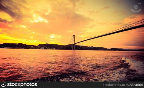 Dramatic sunset cloudy sky above a surface of the sea and long suspension bridge in Bergen, Norway. View from boat