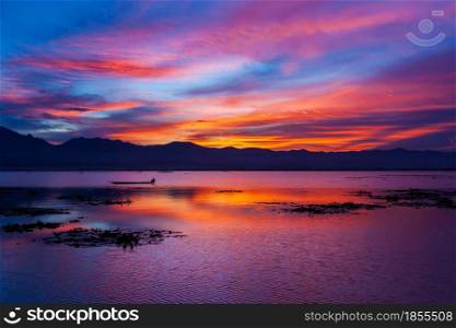 Dramatic sunset sky over the lake and mountains in the backgrounds. Colourful clouds and sky reflection on the lake. Fisherman fishing on the boat. Phayao Lake, Thailand.