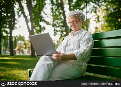 Drandma in glasses using laptop on the bench in summer park. Aged people lifestyle. Pretty grandmother having fun outdoors, old female person on nature. Drandma using laptop on the bench in summer park