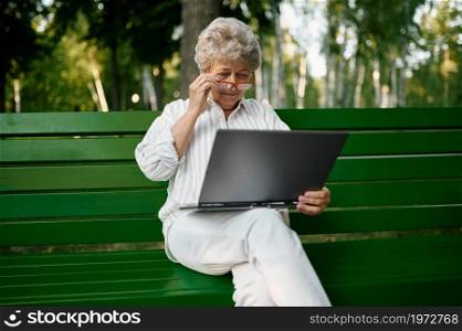 Drandma in glasses using laptop on the bench in summer park. Aged people lifestyle. Pretty grandmother having fun outdoors, old female person on nature. Drandma using laptop on the bench in summer park