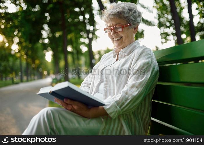 Dranny in glasses reading a book on the bench in summer park. Aged people lifestyle. Pretty grandmother having fun outdoors, old female person on nature. Dranny reading a book on the bench in summer park