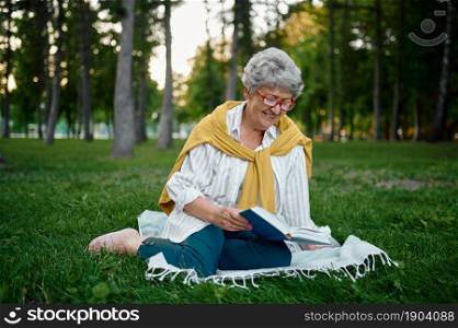 Dranny in glasses reading a book on the grass in summer park. Aged people lifestyle. Pretty grandmother having fun outdoors, old female person on nature. Dranny in glasses reading a book on the grass