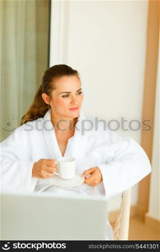 Dreaming woman in bathrobe with laptop on table having cup of coffee&#xA;