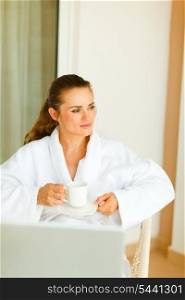 Dreaming woman in bathrobe with laptop on table having cup of coffee&#xA;