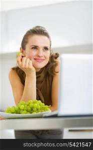 Dreaming young housewife eating grape in kitchen