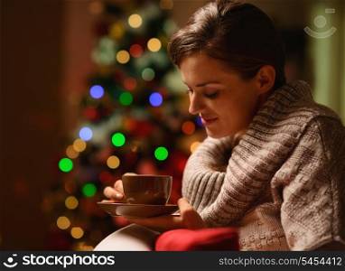 Dreaming young woman sitting chair with hot chocolate in front of Christmas tree