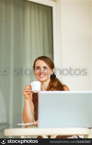 Dreaming young woman sitting on terrace with laptop and cup of coffee