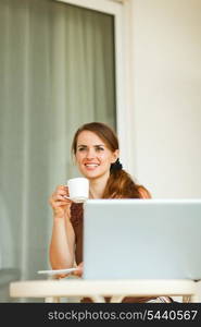 Dreaming young woman sitting on terrace with laptop and cup of coffee