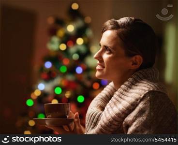 Dreaming young woman with cup of hot beverage in front of christmas lights