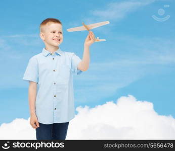 dreams, future, hobby and childhood concept - smiling little boy holding wooden airplane model in his hand over blue sky background