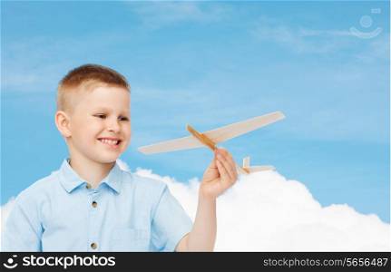 dreams, future, hobby and childhood concept - smiling little boy holding wooden airplane model in his hand over blue sky background