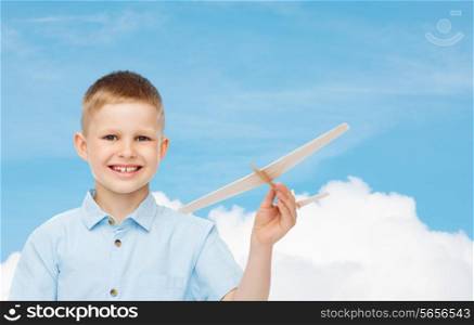 dreams, future, hobby and childhood concept - smiling little boy holding wooden airplane model in his hand over blue sky background