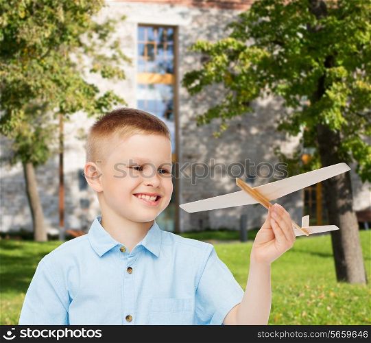 dreams, future, hobby, education and childhood concept - smiling little boy holding wooden airplane model in his hand over yard background