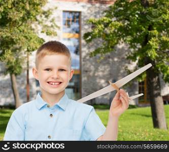 dreams, future, hobby, education and childhood concept - smiling little boy holding wooden airplane model in his hand over yard background