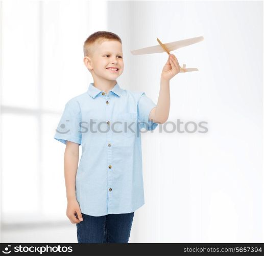dreams, future, hobby, home and childhood concept - smiling little boy holding wooden airplane model in his hand over white room background