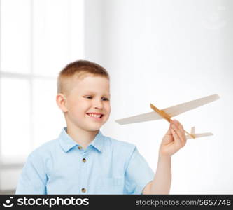 dreams, future, hobby, home and childhood concept - smiling little boy holding wooden airplane model in his hand over white room background