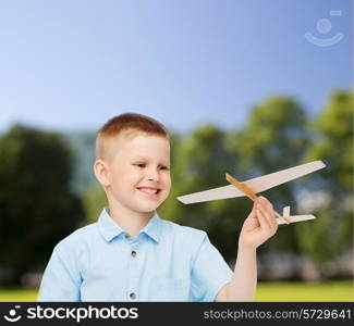 dreams, future, hobby, nature and childhood concept - smiling little boy holding wooden airplane model in his hand over park background