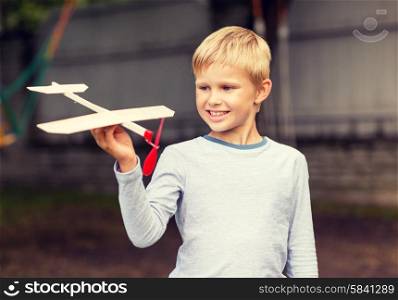 dreams, future, hobby, people and childhood concept - smiling little boy holding wooden airplane model in his hand outdoors