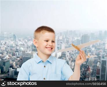 dreams, future, hobby, urban life and childhood concept - smiling little boy holding wooden airplane model in his hand over city background