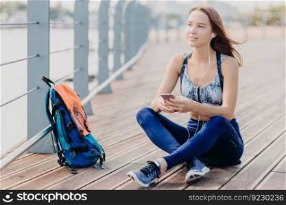 Dreamy attractive female wears top and leggings, thinks about something while listens music, holds modern cell phone, bag stands near, breathes fresh air in morning. Recreation and lifestyle concept