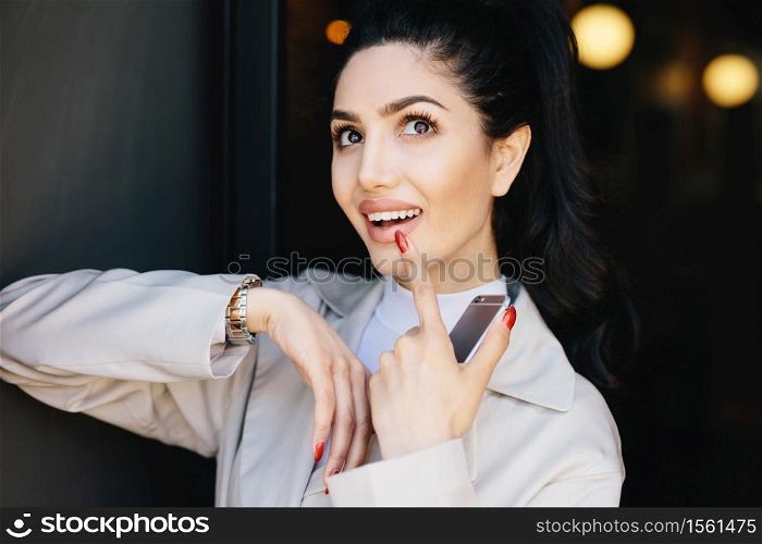Dreamy beautiful brunette model with curling long eyelashes, dark bright eyes and red manicure dressed in white jacket holding her finger on lips looking up while dreaming about something pleasant