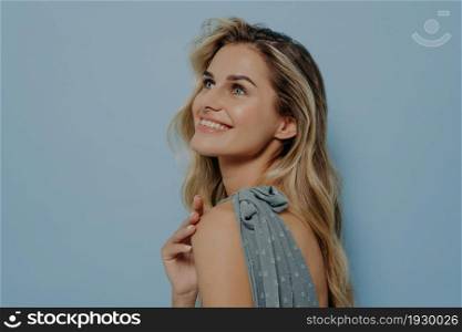 Dreamy blonde woman in blue dress thinking about pleasant things, looking upwards with sincere smile, dreaming of something, standing isolated in front of blue background. Dreamy blonde woman dreaming and looking upwards