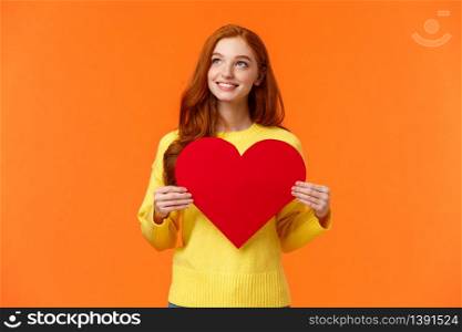Dreamy cute redhead girl waiting for her prince, planning perfect velantines day romantic date, holding large heart sign and looking upper left corner with tender happy smile, orange background.. Dreamy cute redhead girl waiting for her prince, planning perfect velantines day romantic date, holding large heart sign and looking upper left corner with tender happy smile, orange background