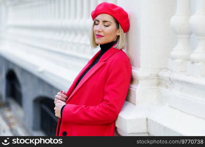 Dreamy female with blond hair in red clothes with beret standing with closed eyes near white fence on street of city. Charming woman in red outfit near fence