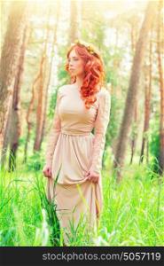 Dreamy girl standing in a forest on sunny spring day, wearing long elegant dress and flower wreath on curly red hair, fashion look