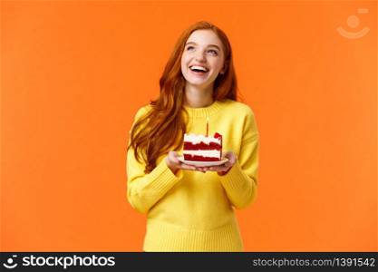 Dreamy happy girl celebrating birthday, having fun, laughing and gazing upper left corner thoughtful, making wish as blowing out candle on delicious, tasty piece b-day cake, orange background.. Dreamy happy girl celebrating birthday, having fun, laughing and gazing upper left corner thoughtful, making wish as blowing out candle on delicious, tasty piece b-day cake, orange background