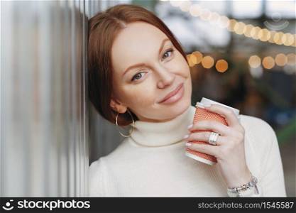 Dreamy pensive Caucasian woman with make up, dressed in casual outfit, leans on wall, holds takeaway coffee, remembers pleasant moments during outdoor stroll, has appealing look. Drink concept
