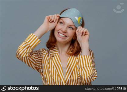 Dreamy redhaired European woman looks gladfully aside, smiles broadly, wears casual domestic clothes and sleepmask, isolated over grey background, imagines something pleasant. Good morning concept
