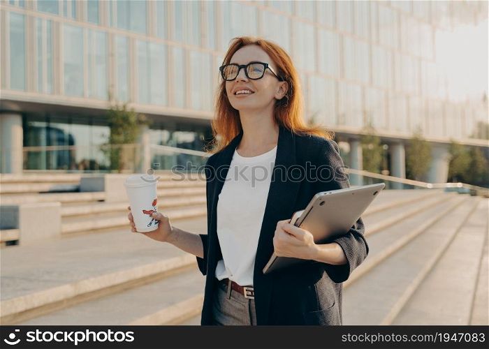 Dreamy redhead woman in formal clothes carries notepad tablet disposable cup of coffee has walk outdoor during daytime poses near office building with cheerful expression going to have job interview. Dreamy redhead woman in formal clothes carries notepad tablet disposable cup of coffee