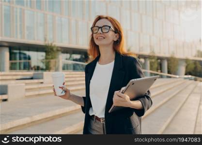 Dreamy redhead woman in formal clothes carries notepad tablet disposable cup of coffee has walk outdoor during daytime poses near office building with cheerful expression going to have job interview. Dreamy redhead woman in formal clothes carries notepad tablet disposable cup of coffee